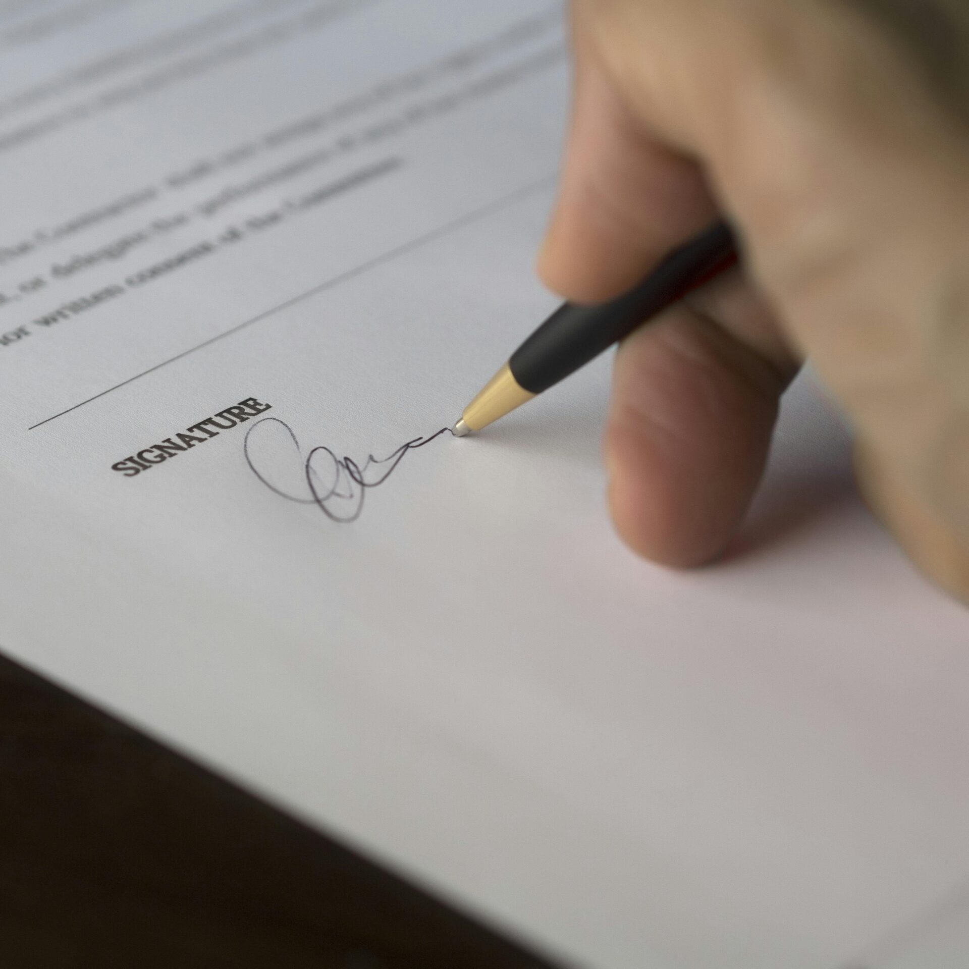 A hand signs a formal contract with a pen on a wooden desk.