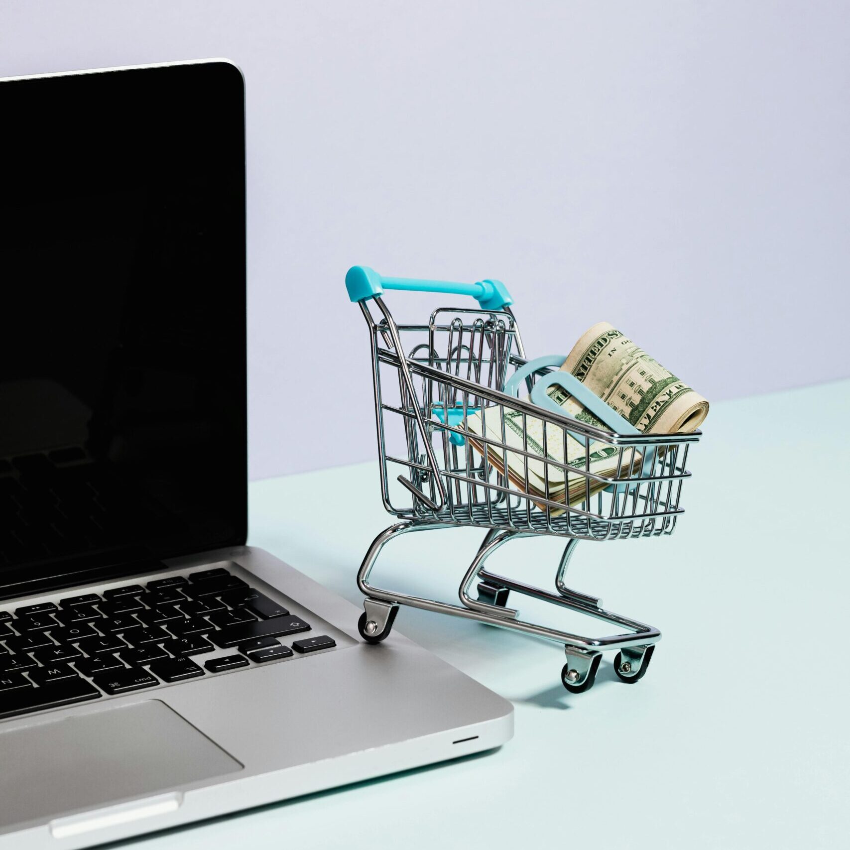 Mini shopping cart with rolled cash next to a laptop, representing online shopping.